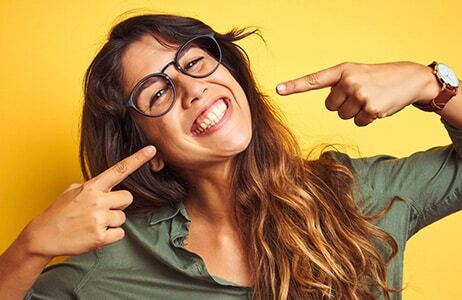 Smiling woman wearing glasses pointing at her teeth against yellow background.