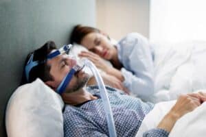 Man wearing CPAP mask while sleeping next to woman, illustrating sleep apnea treatment.