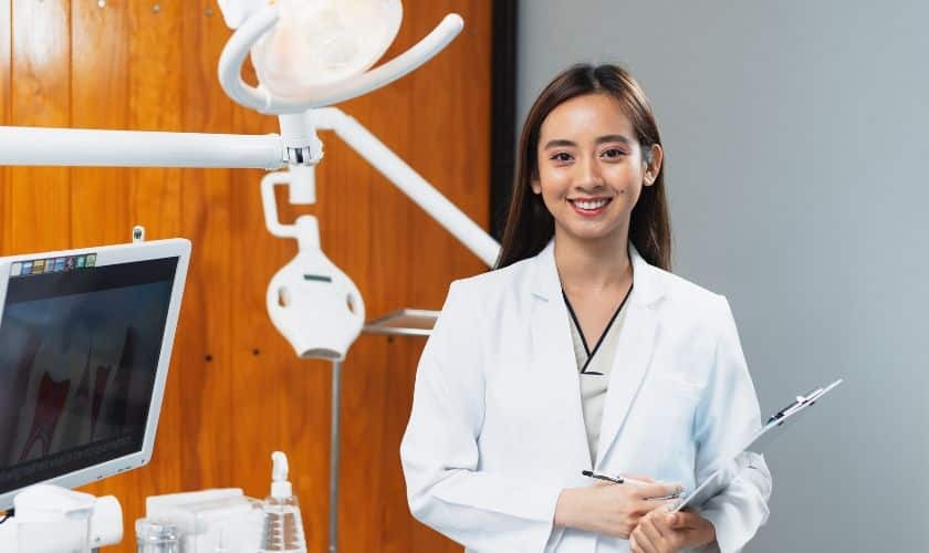 Smiling female dentist in white coat holding tablet in modern dental office with surgical light.