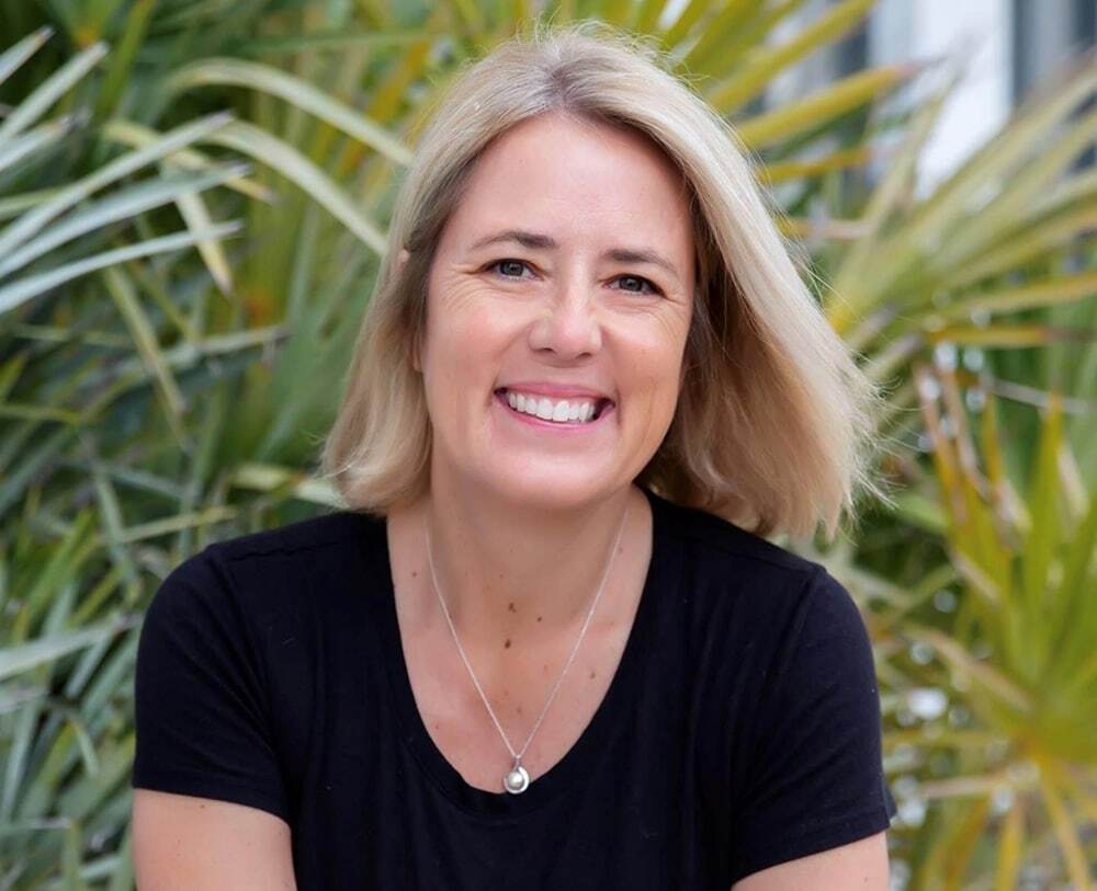Smiling woman with blonde hair wearing a black shirt in front of palm plants.