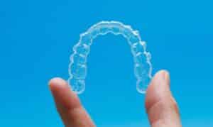 Clear Invisalign aligner tray held between fingers against blue background.