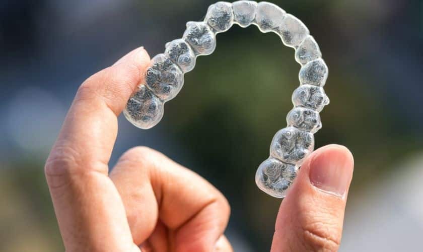 Clear Invisalign aligner held between fingers, showing transparent plastic tray with multiple aligners.