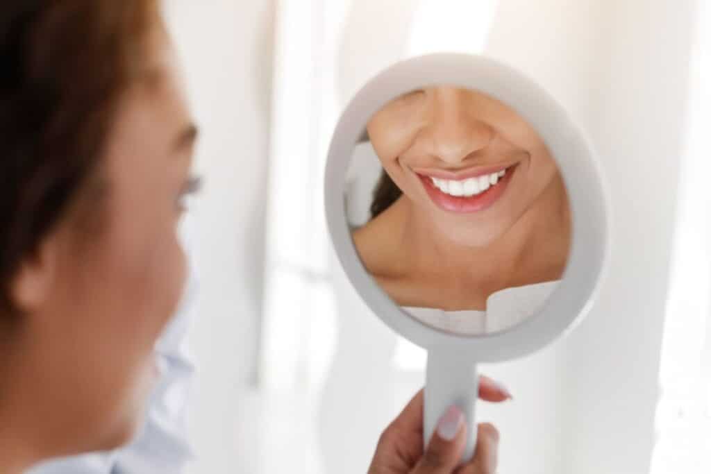 Woman smiling in handheld mirror, displaying bright white teeth and healthy smile.