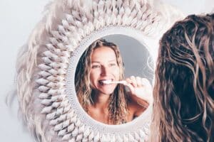 Woman brushing teeth while looking in a decorative mirror with feathered frame.