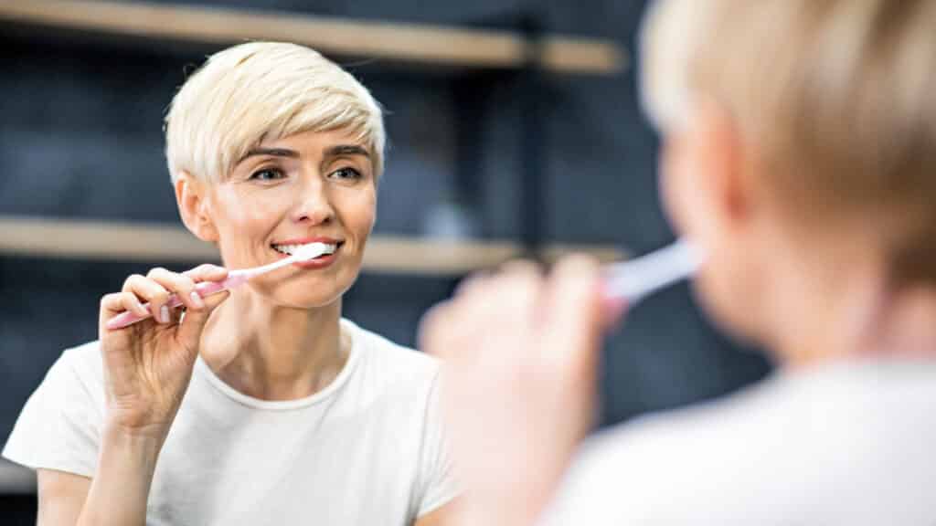 Woman brushing her teeth in bathroom mirror, demonstrating daily dental hygiene habits.