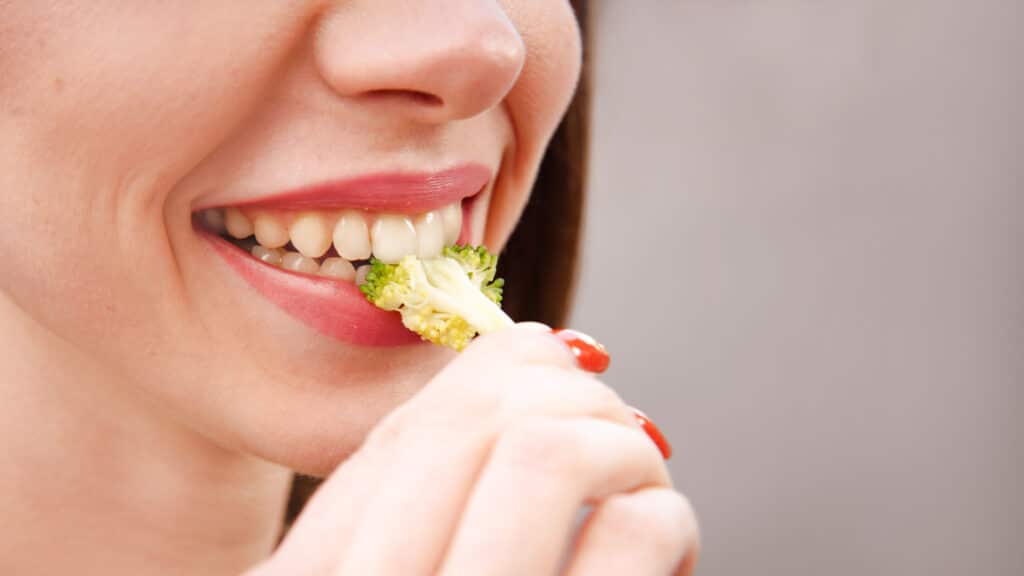 Woman smiling while eating fresh broccoli, demonstrating healthy eating habits for dental health.