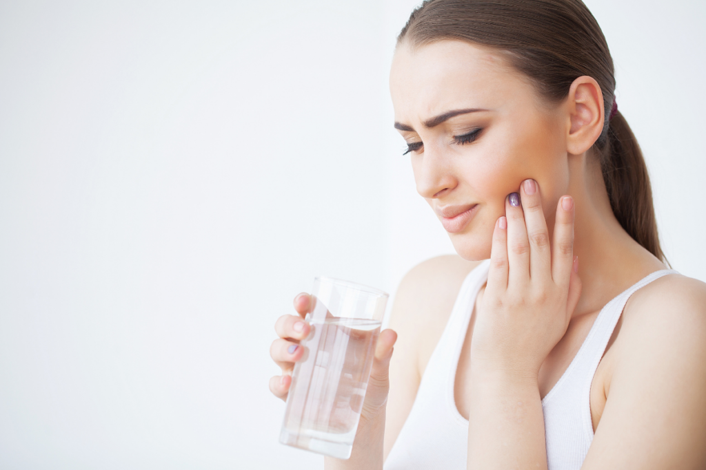 Woman experiencing tooth pain, holding a glass of water and touching her cheek.